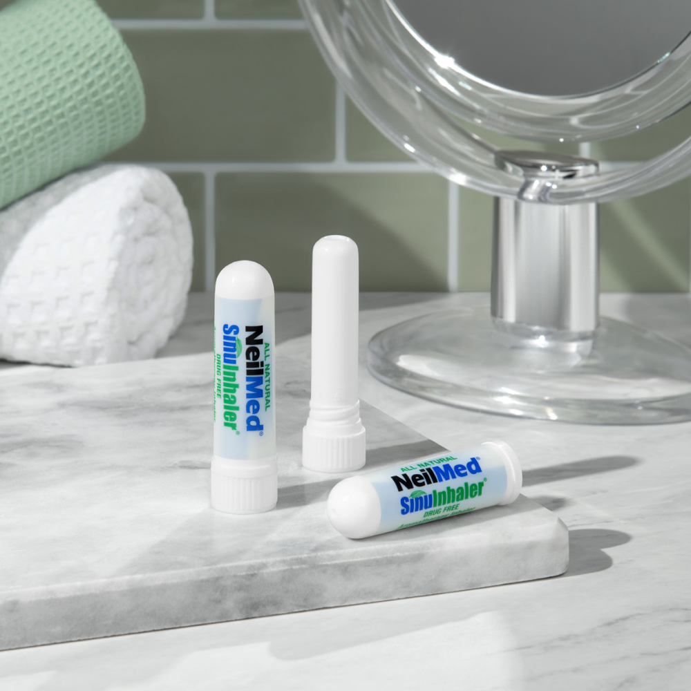 NeilMed SinuInhaler sticks displayed on a bathroom countertop next to a mirror and towels, with one inhaler open and two closed.