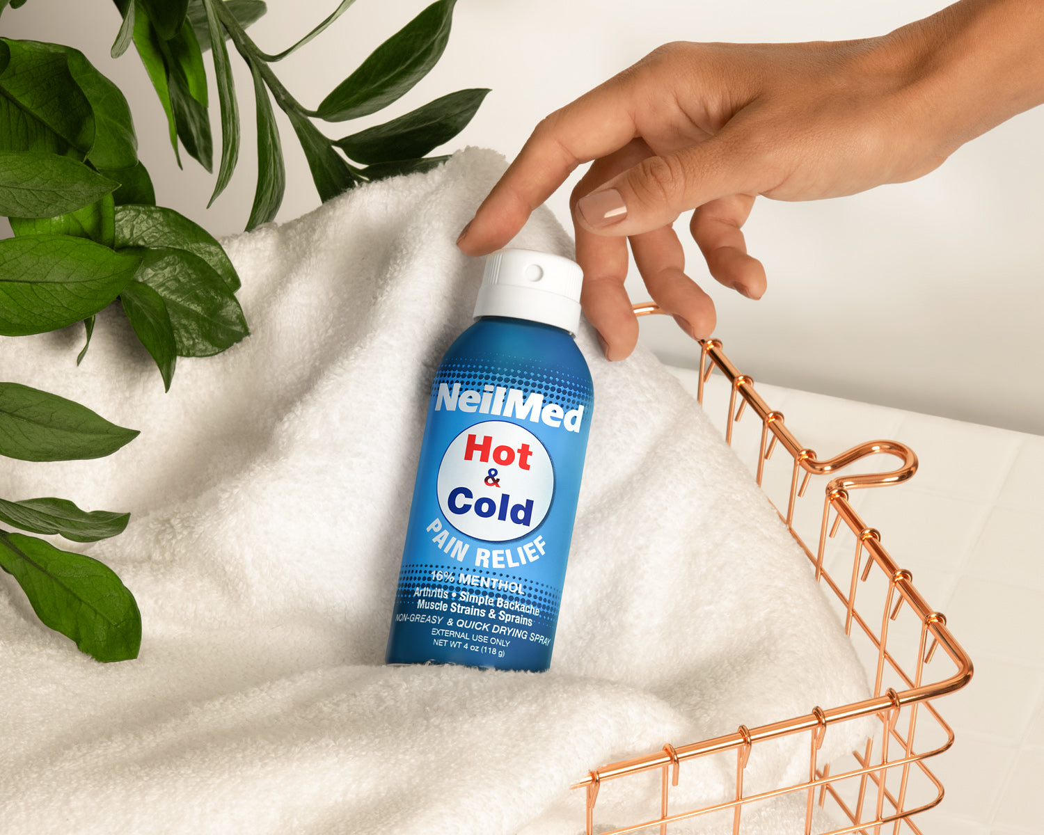 Hand reaching for a NeilMed Hot & Cold Pain Reliever spray bottle on a white surface with green leaves in the background.